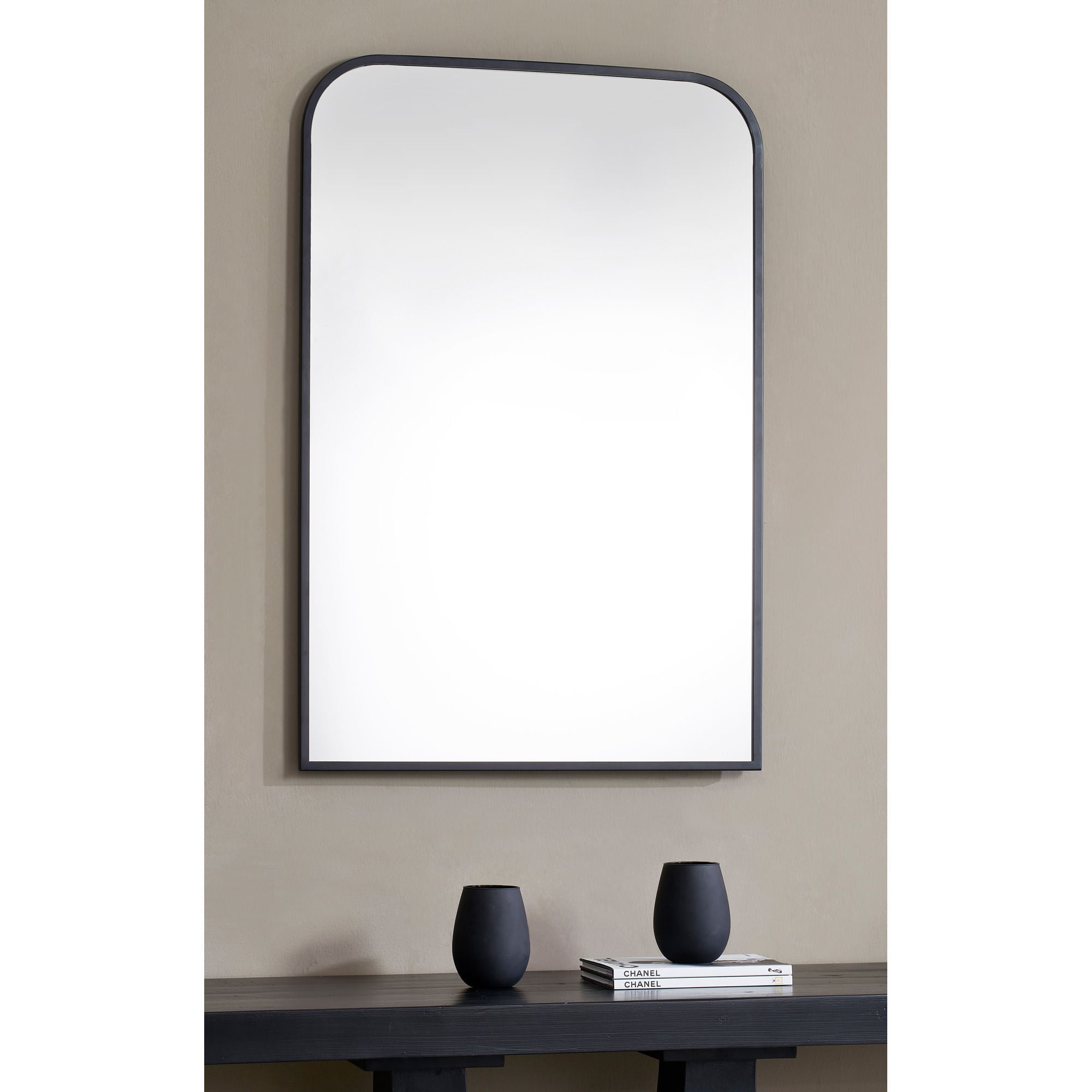 Ammar mirror with a sleek black frame and rounded top, mounted on a beige wall above a dark wooden table with black vases.