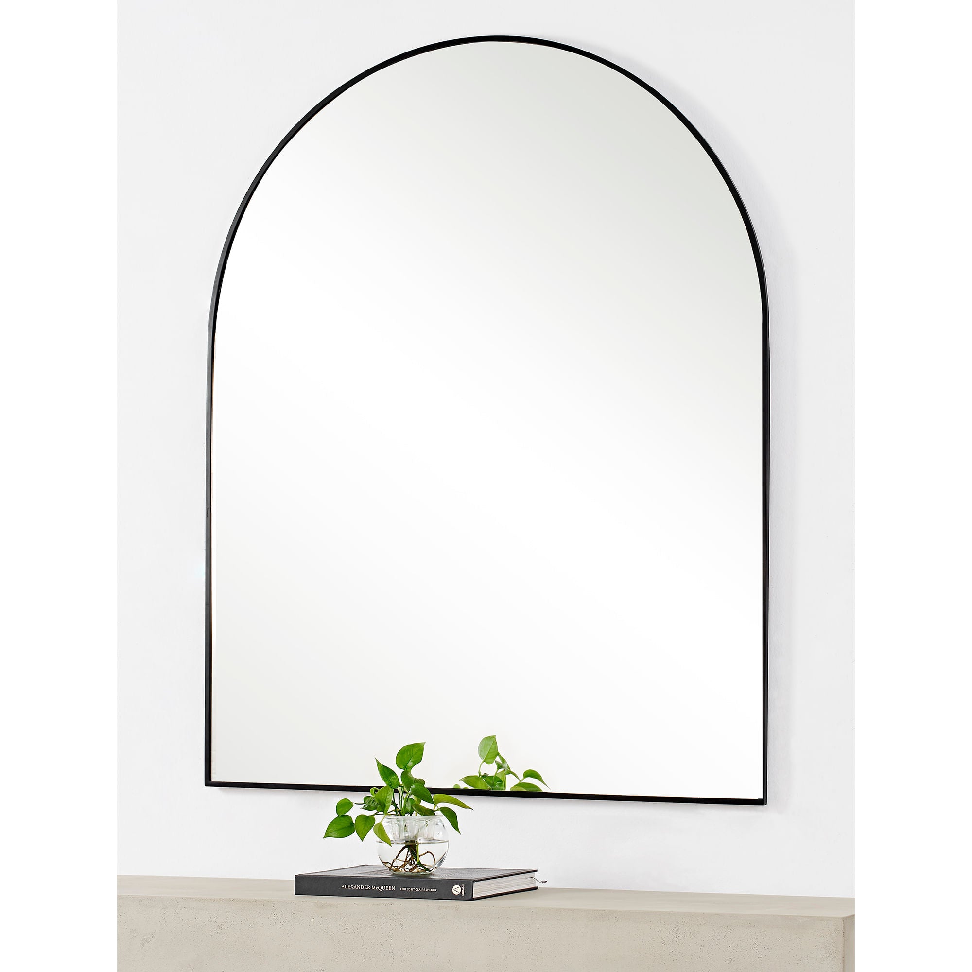 Lamia mirror with an arched top, featuring a sleek black frame, displayed above a concrete surface with a small plant and books.