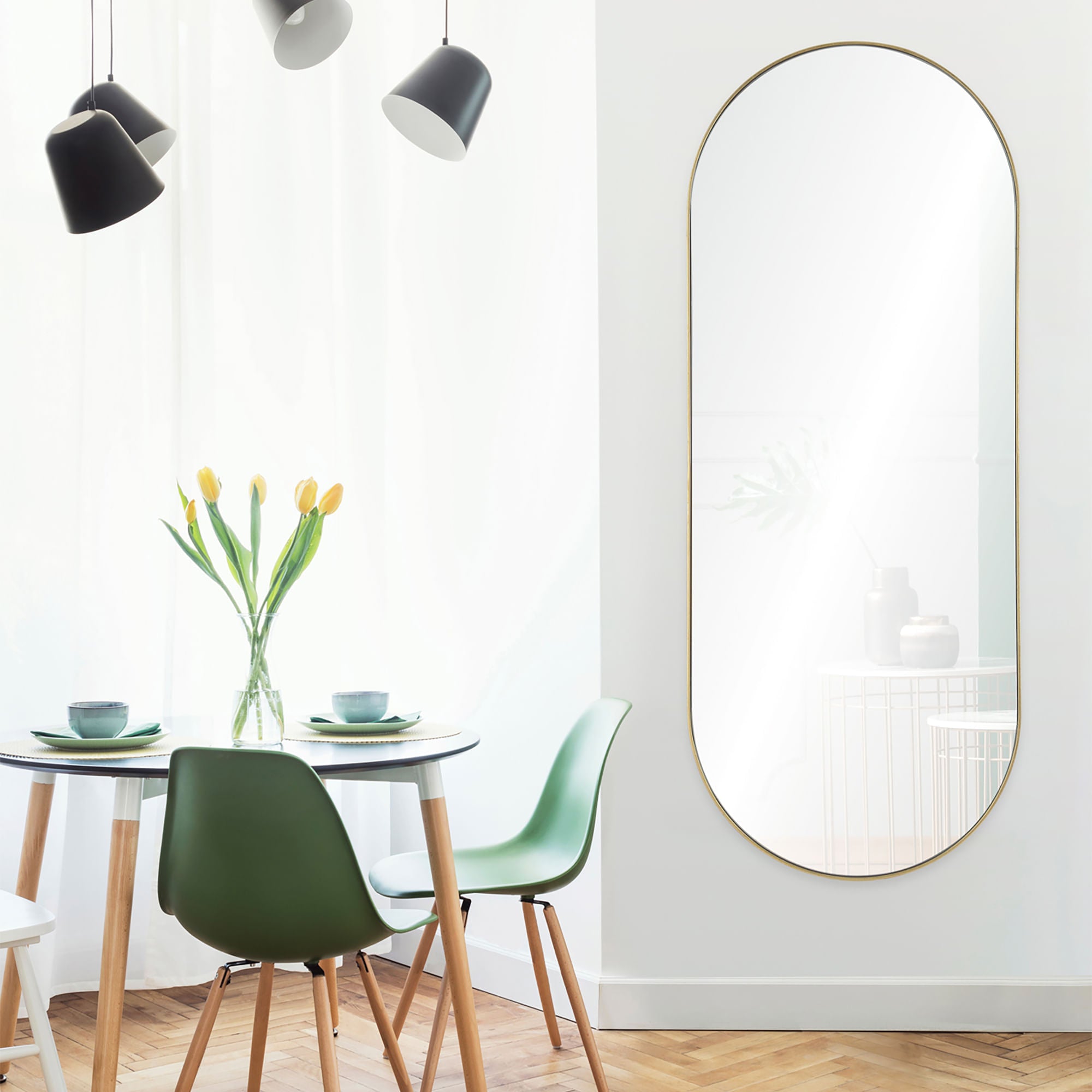 Marius mirror with a gold frame, displayed on a white wall next to a dining table with green chairs and yellow tulips.