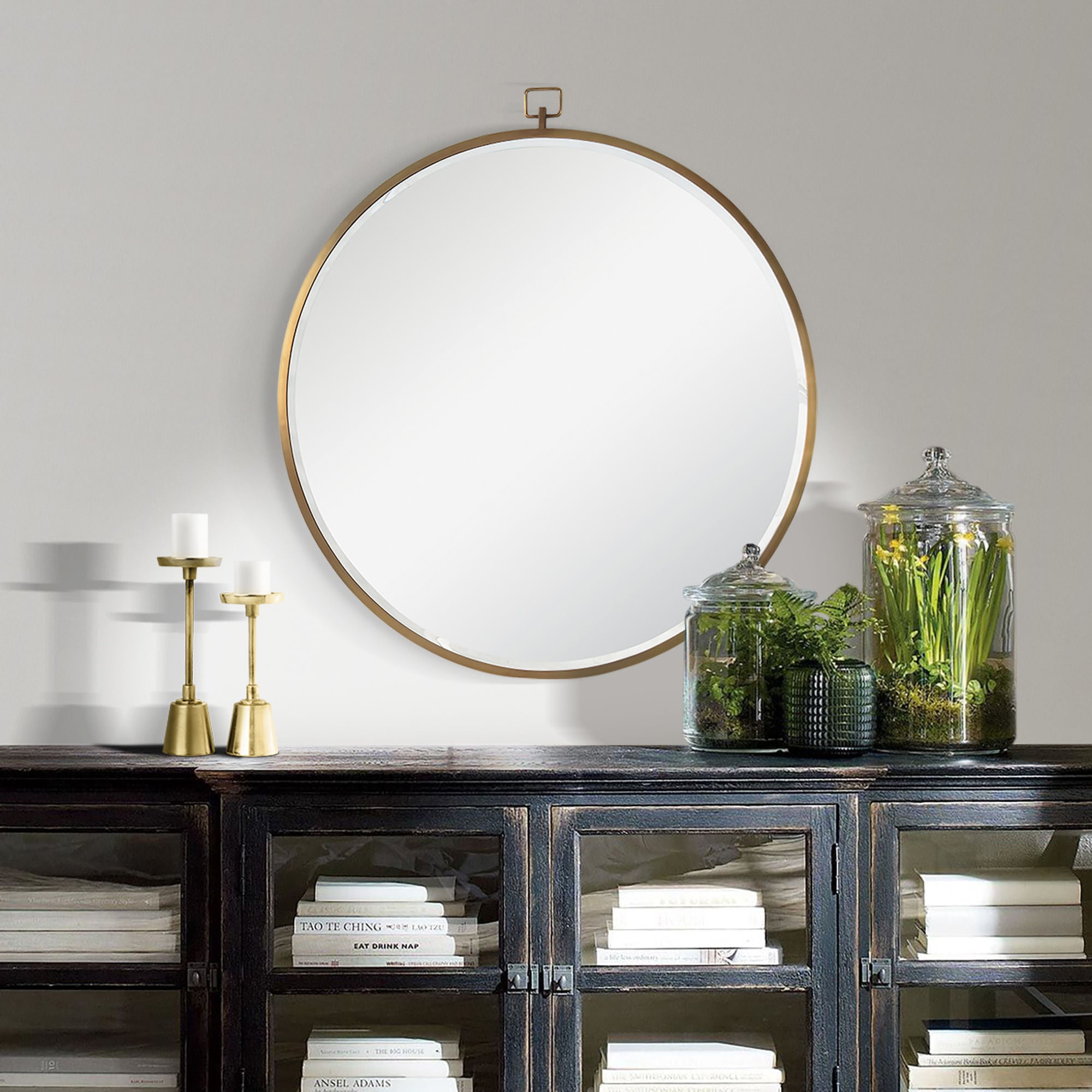 Azam round mirror with a gold frame, hanging above a dark wood cabinet with candles and glass jars of plants.