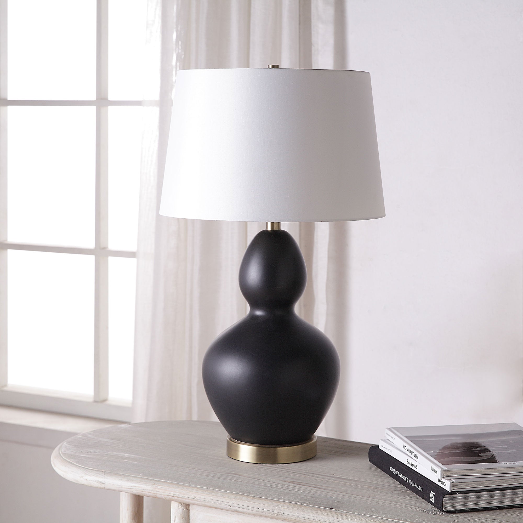 Kamilia Table Lamp placed on a table beside books, featuring a black ceramic body and white shade.