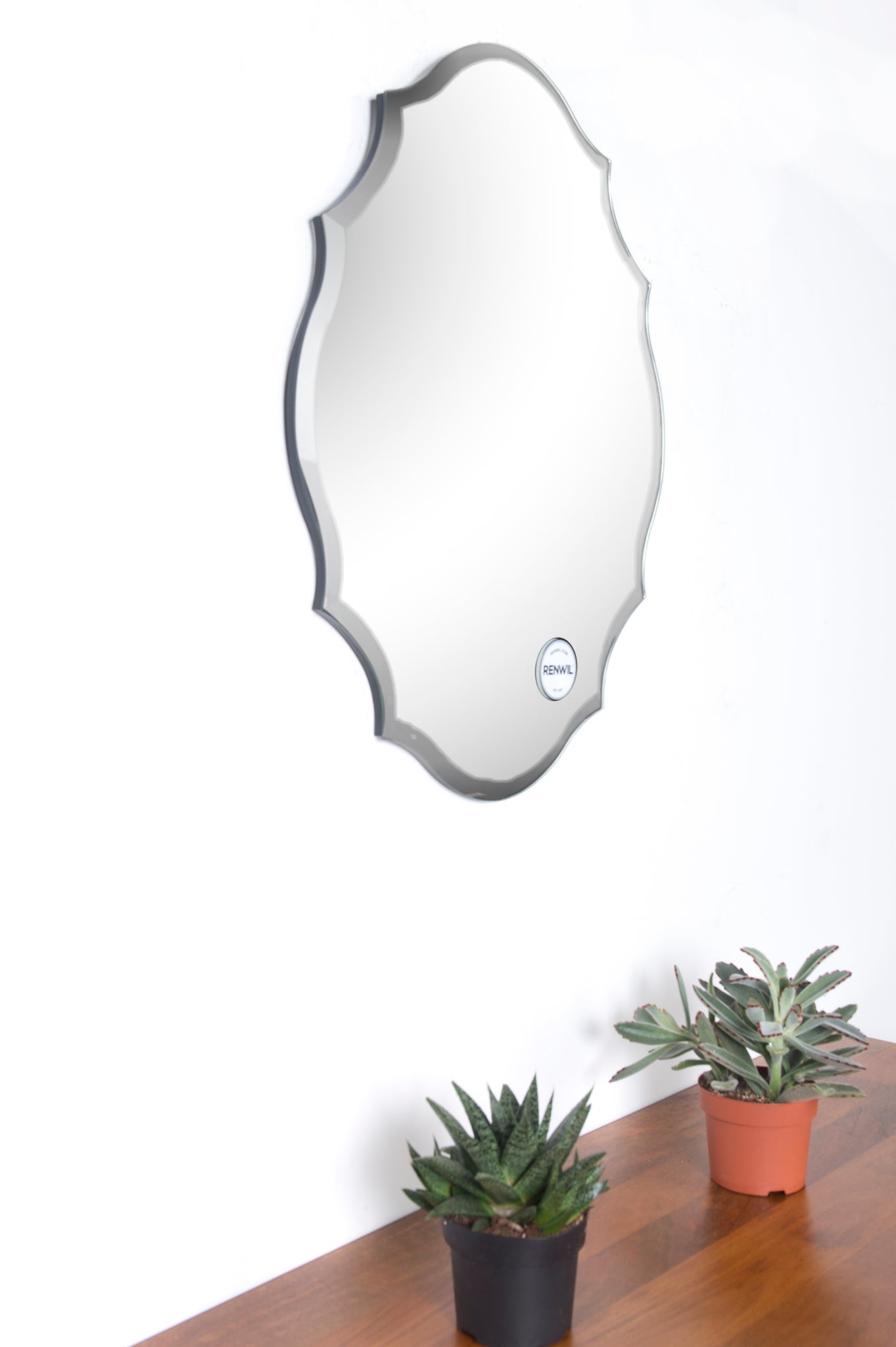 Emma mirror by Renwil mounted on a wall, surrounded by potted plants, scalloped edges visible.