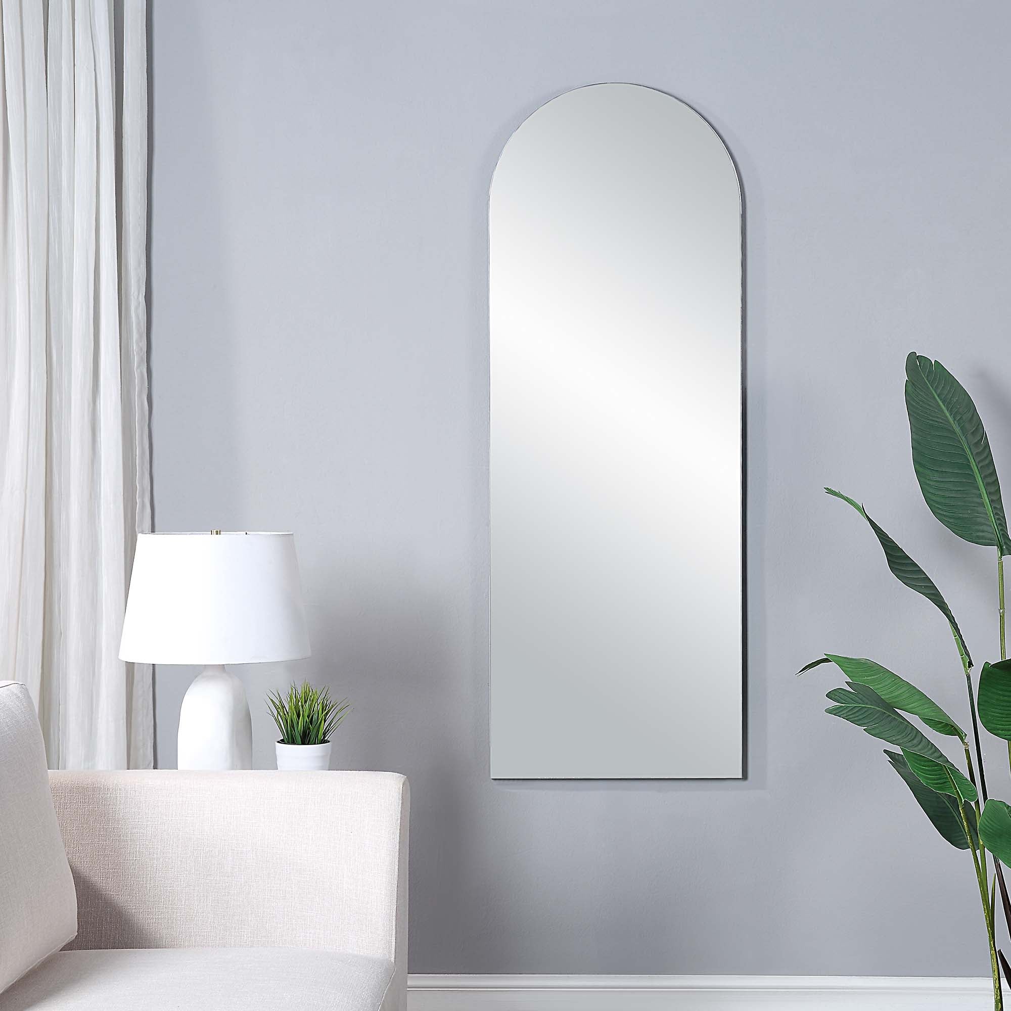 Mandra mirror with an arched top, mounted on a light gray wall, beside a white lamp and green plant.