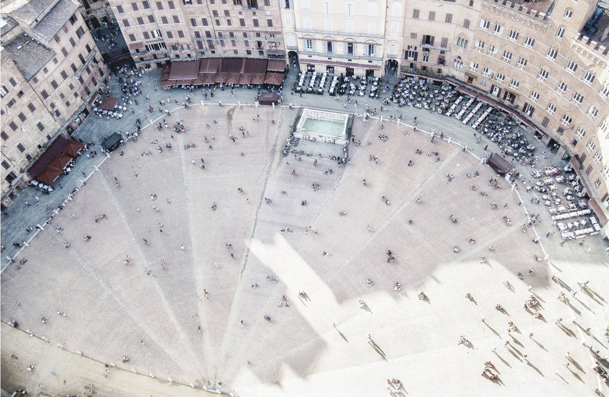 Piazza De Campo, an aerial view of a bustling square with people, cafes, and a central fountain, featuring sandy tones.