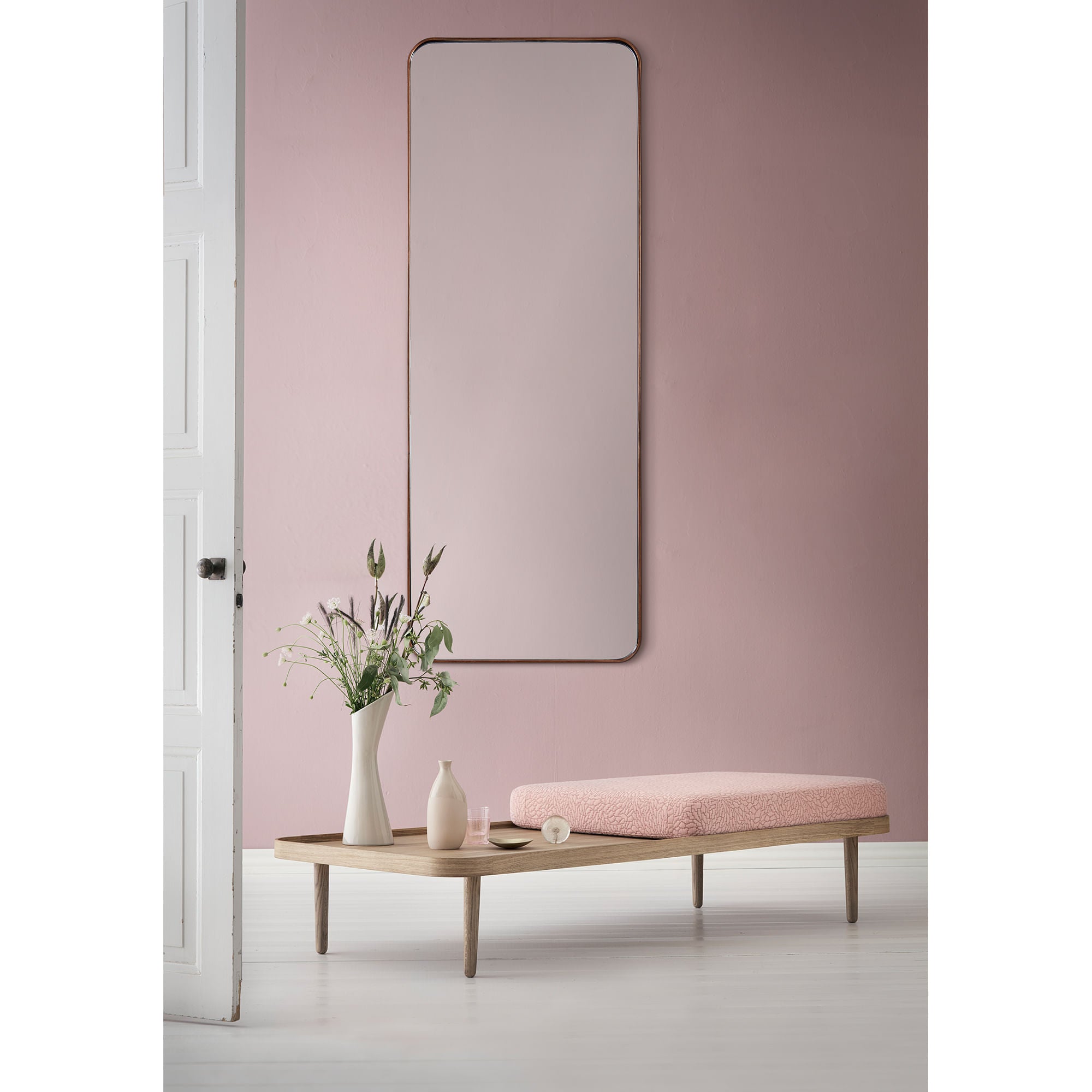 Phiale mirror mounted on a pink wall above a wooden table with decorative vases.