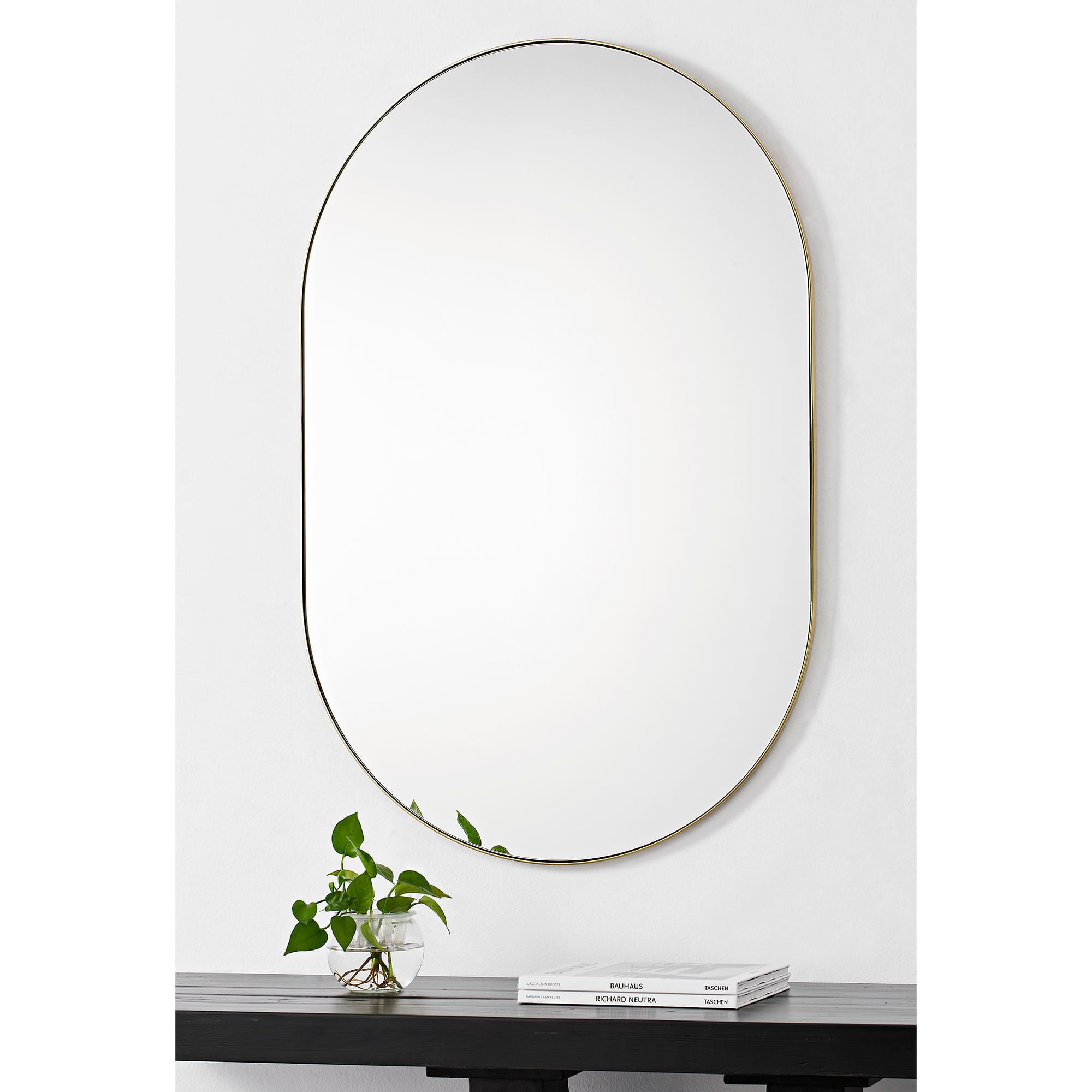 Beckah mirror with an oval shape and thin gold frame, mounted on a white wall above a black table with a plant.