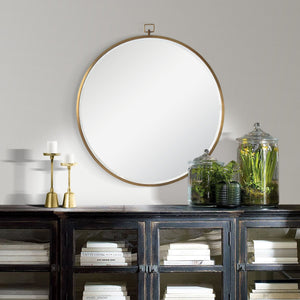 Azam round mirror with a gold frame, hanging above a dark wood cabinet with candles and glass jars of plants.