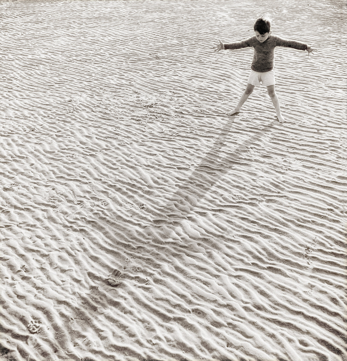 His Shadow, a monochrome wall art featuring a child playing on textured sand, casting a long shadow.
