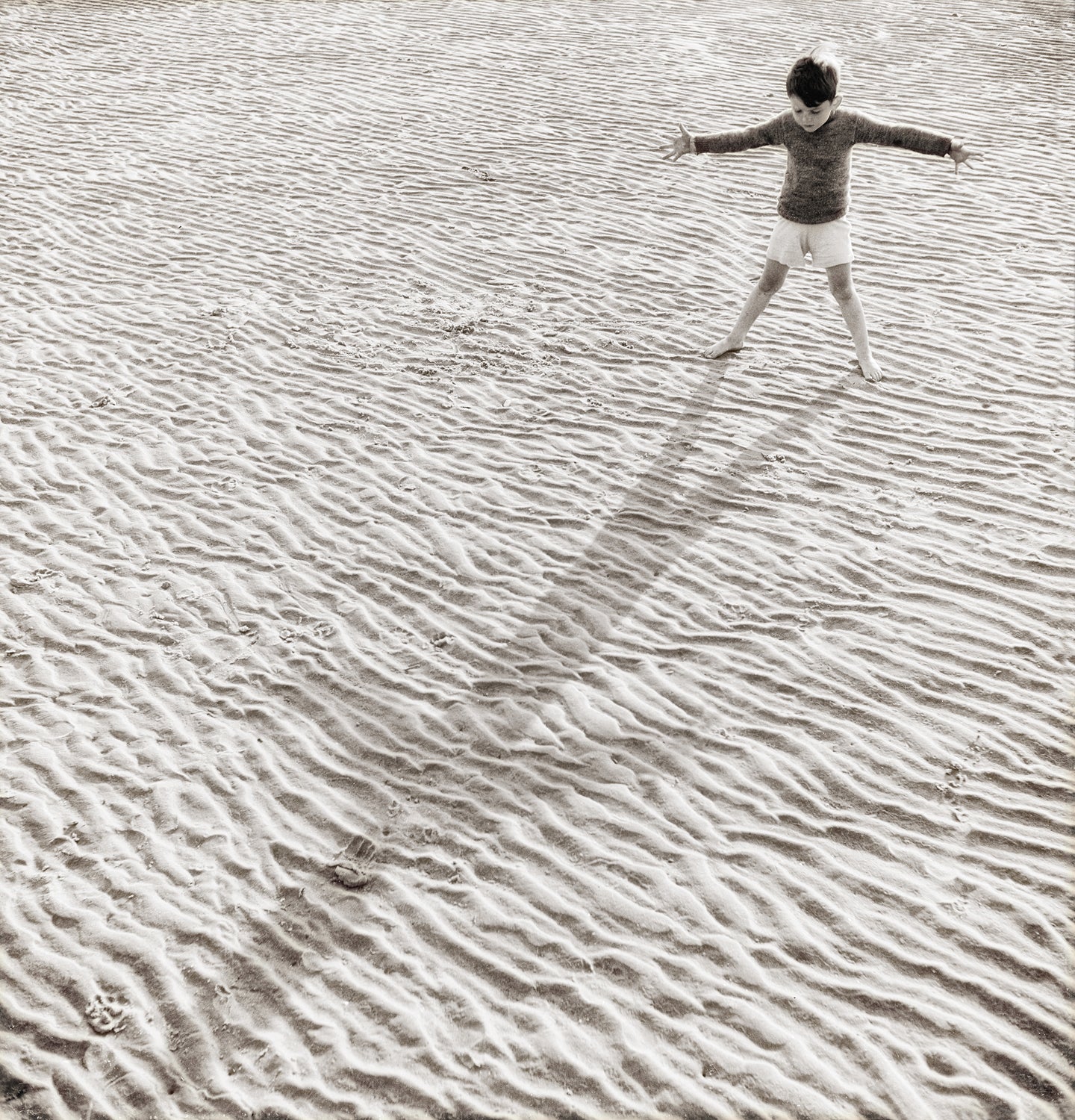 His Shadow, a monochrome wall art featuring a child playing on textured sand, casting a long shadow.
