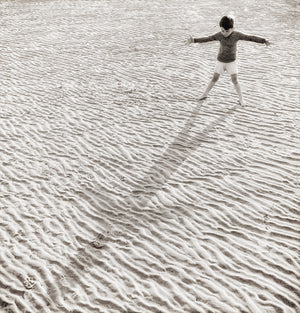His Shadow, a monochrome wall art featuring a child playing on textured sand, casting a long shadow.