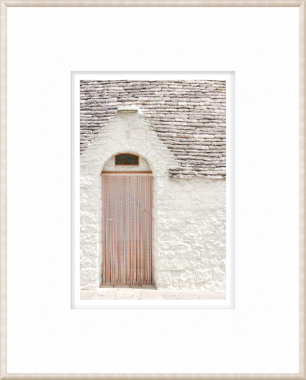 Framed wall art of a white stone wall with a wooden door and hanging strands in Alberobello, Italy.