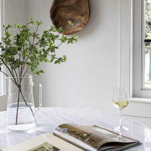 Clear vase with greenery, open book, and glass of white wine on a marble surface.