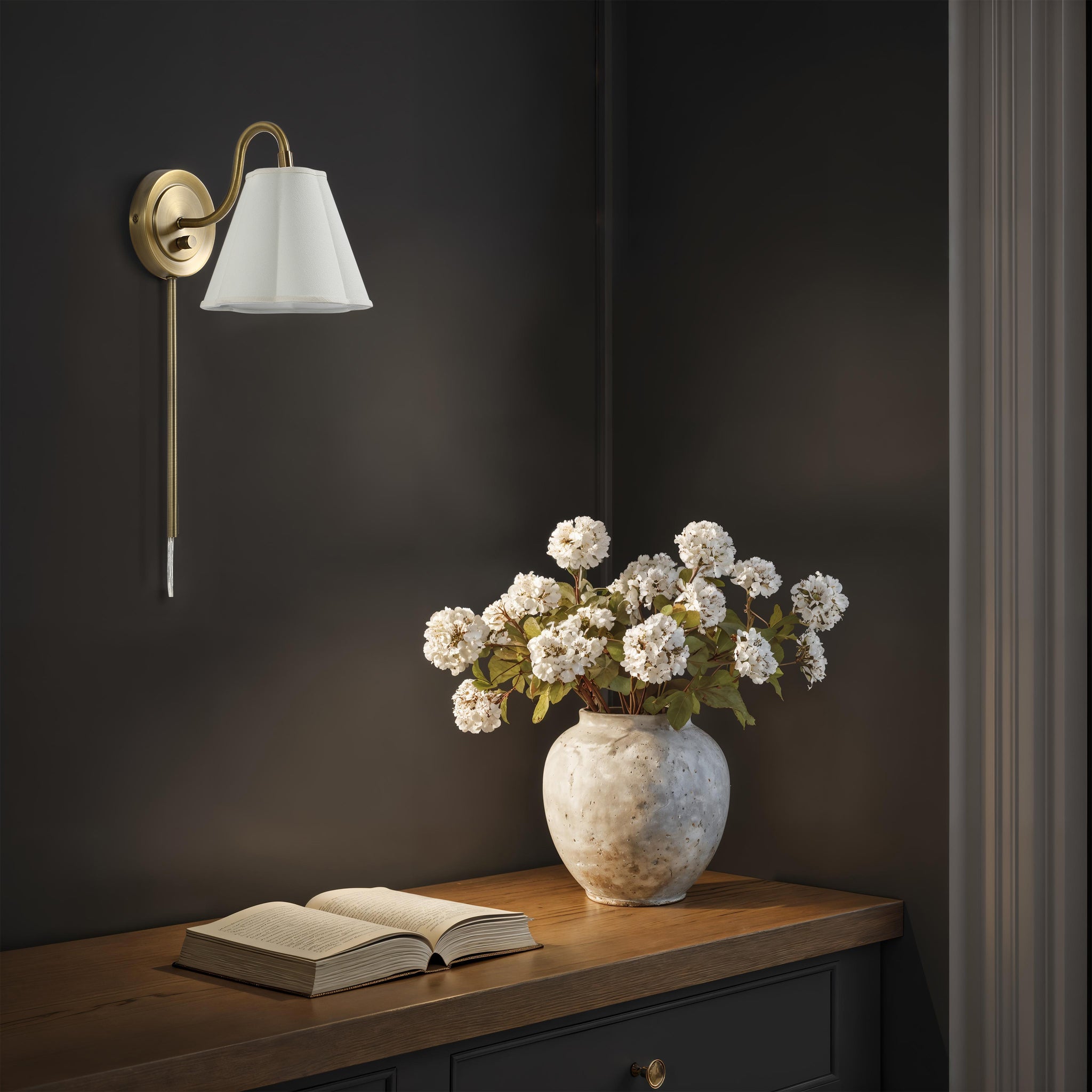 Arielle wall sconce illuminating a dark wall, paired with a vase of flowers and an open book.