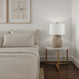 Optima table lamp on a bedside table next to a bed with beige linens and pillows.