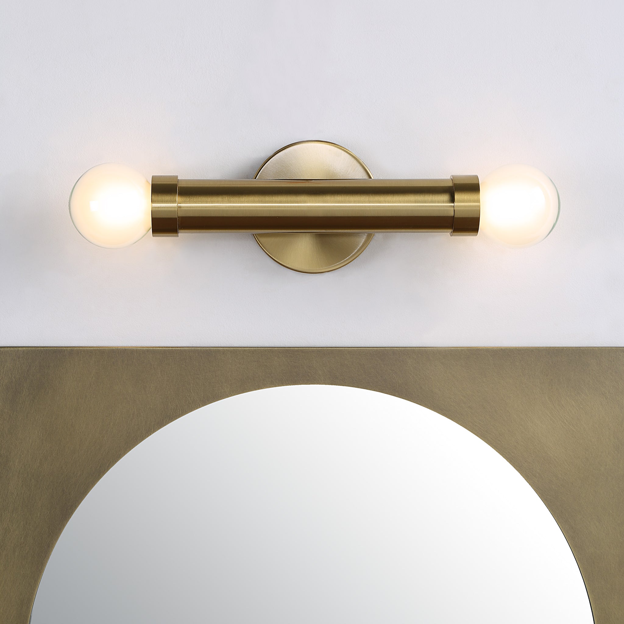 Dasia Wall Sconce in brushed brass above a round mirror, featuring two lit globe bulbs.