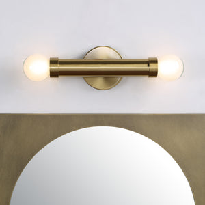 Dasia Wall Sconce in brushed brass above a round mirror, featuring two lit globe bulbs.