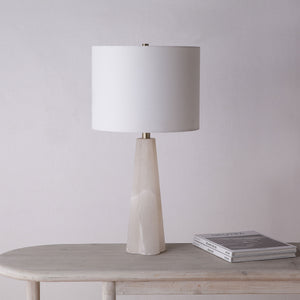 Rima Table Lamp on a wooden table, featuring a white shade and beige stone base against a neutral wall.