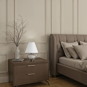 Monumento Table Lamp on a nightstand beside a tufted bed, with decorative pillows and a vase.