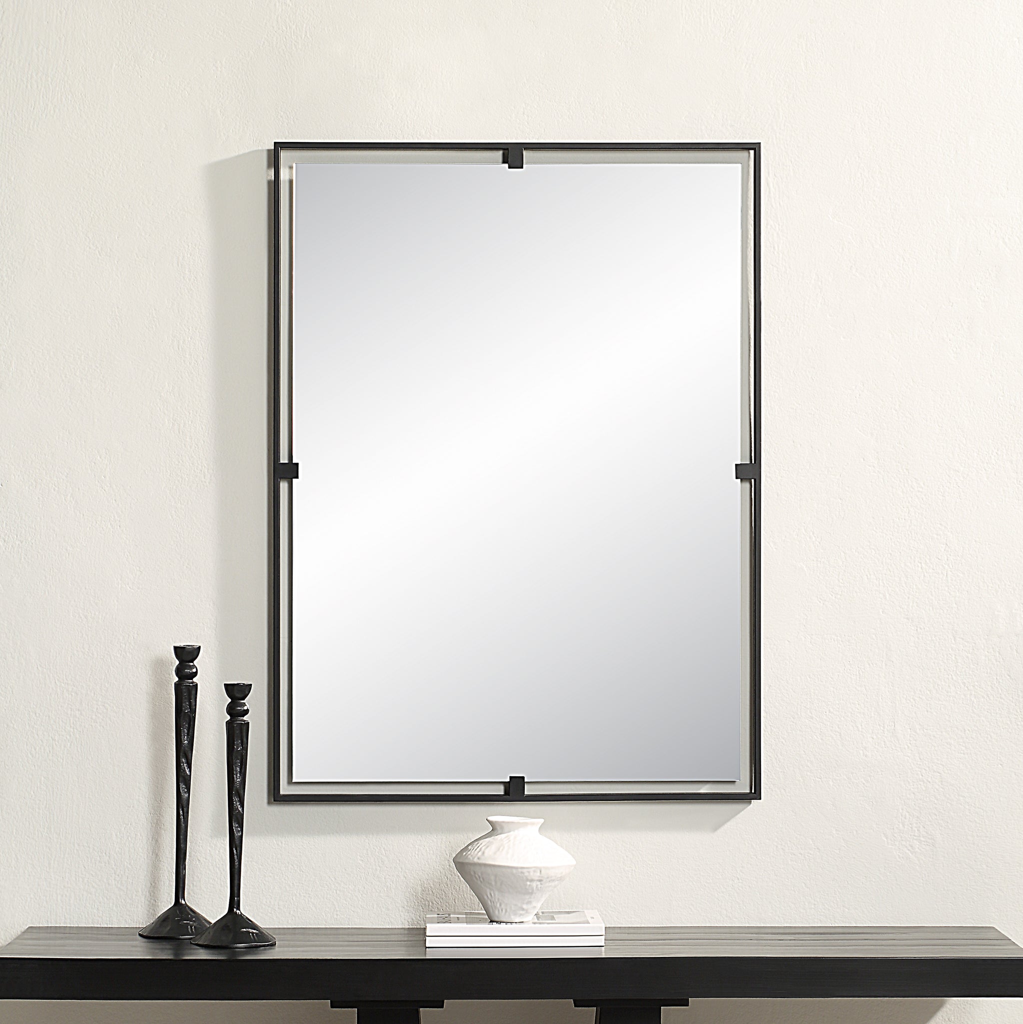 Marco Wall Mirror with a sleek black metal frame, reflecting light, mounted above a dark wood table with decorative items.