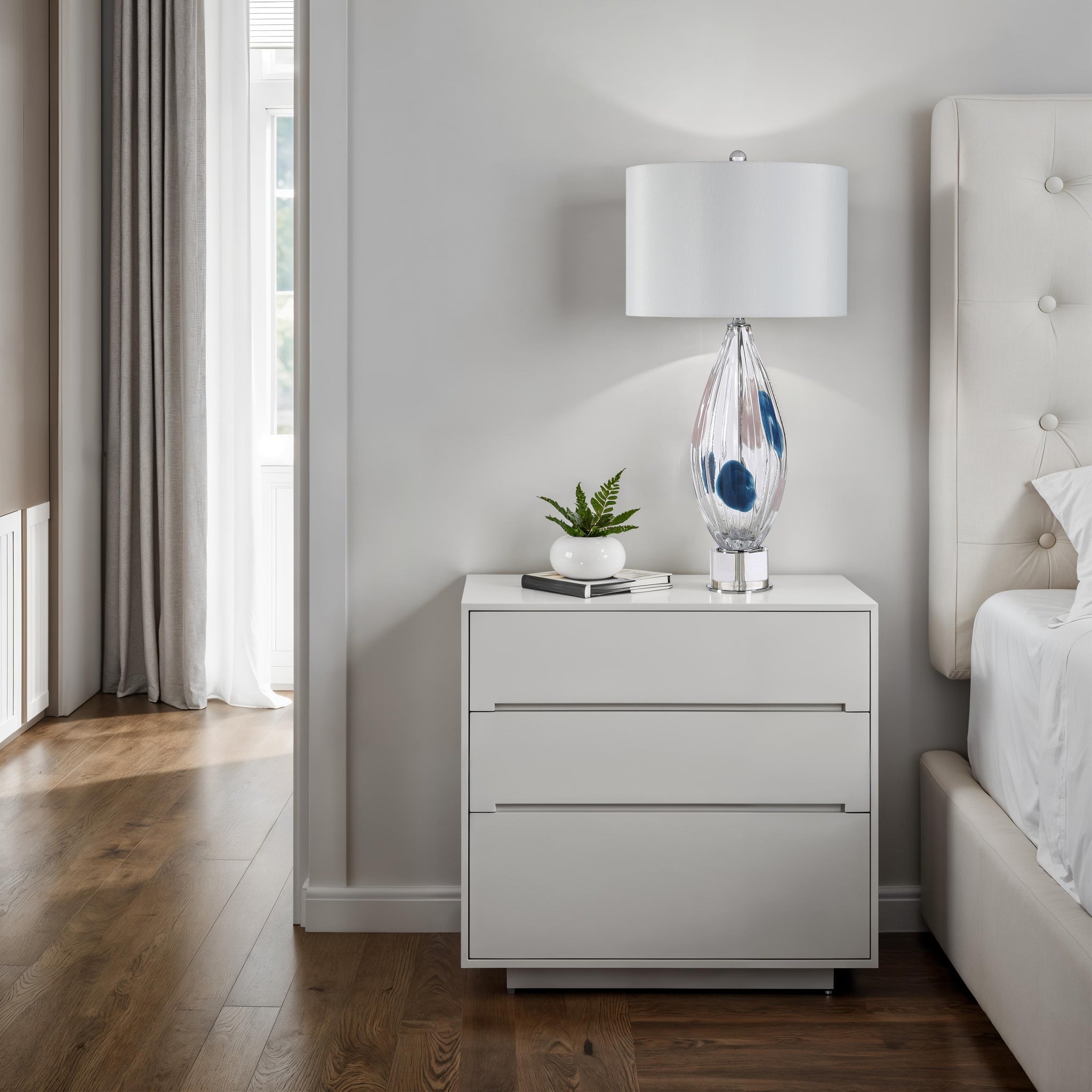 Luxor Table Lamp on a modern nightstand in a bedroom, featuring a clear glass base and white shade