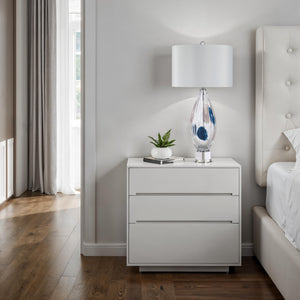 Luxor Table Lamp on a modern nightstand in a bedroom, featuring a clear glass base and white shade