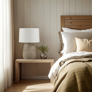 Optima table lamp on a wooden nightstand beside a bed with neutral bedding and a vase.