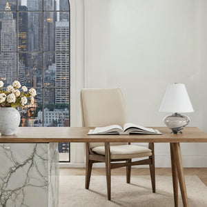 Monumento Table Lamp on a wooden desk with an open book, overlooking a city skyline through a window.