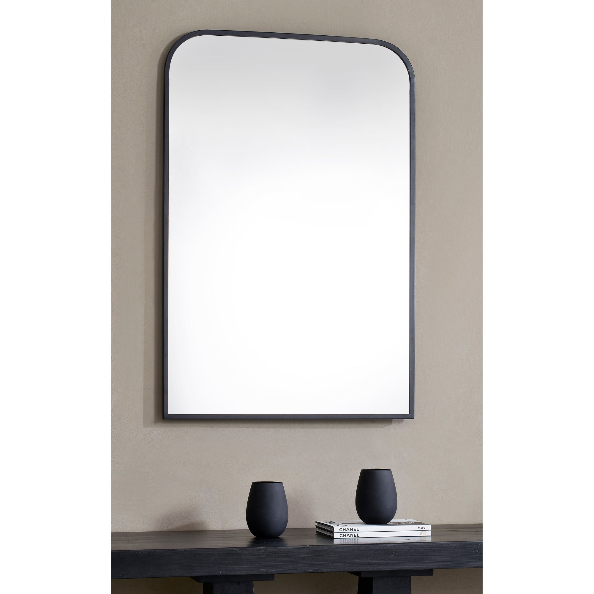 Jackline Wall Mirror displayed on a wall above a table with black vases and books.