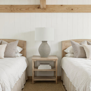 Delfino table lamp on a wooden nightstand between two beds in a bright room.