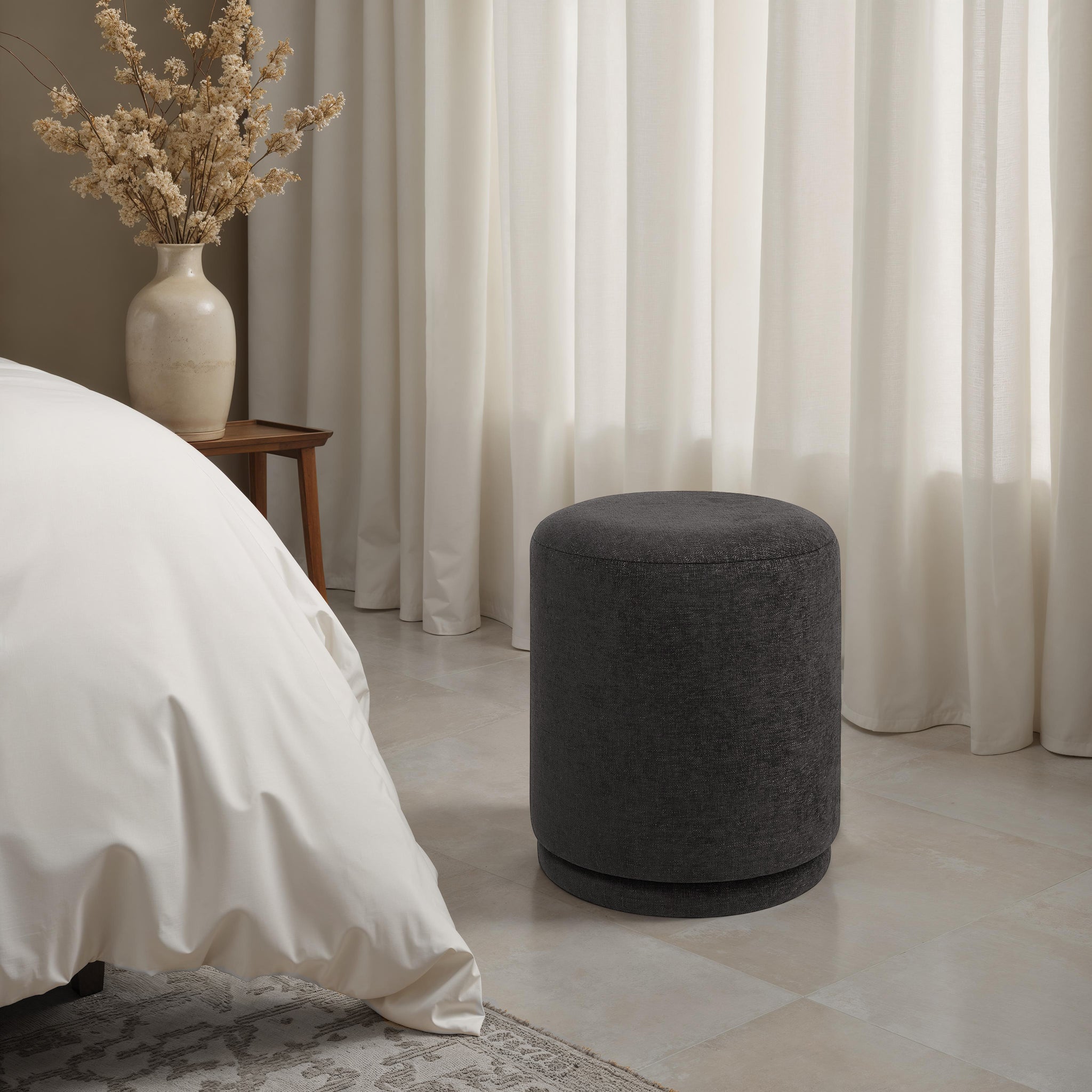 Hevea stool in dark gray placed beside a bed, with curtains and a vase in the background.