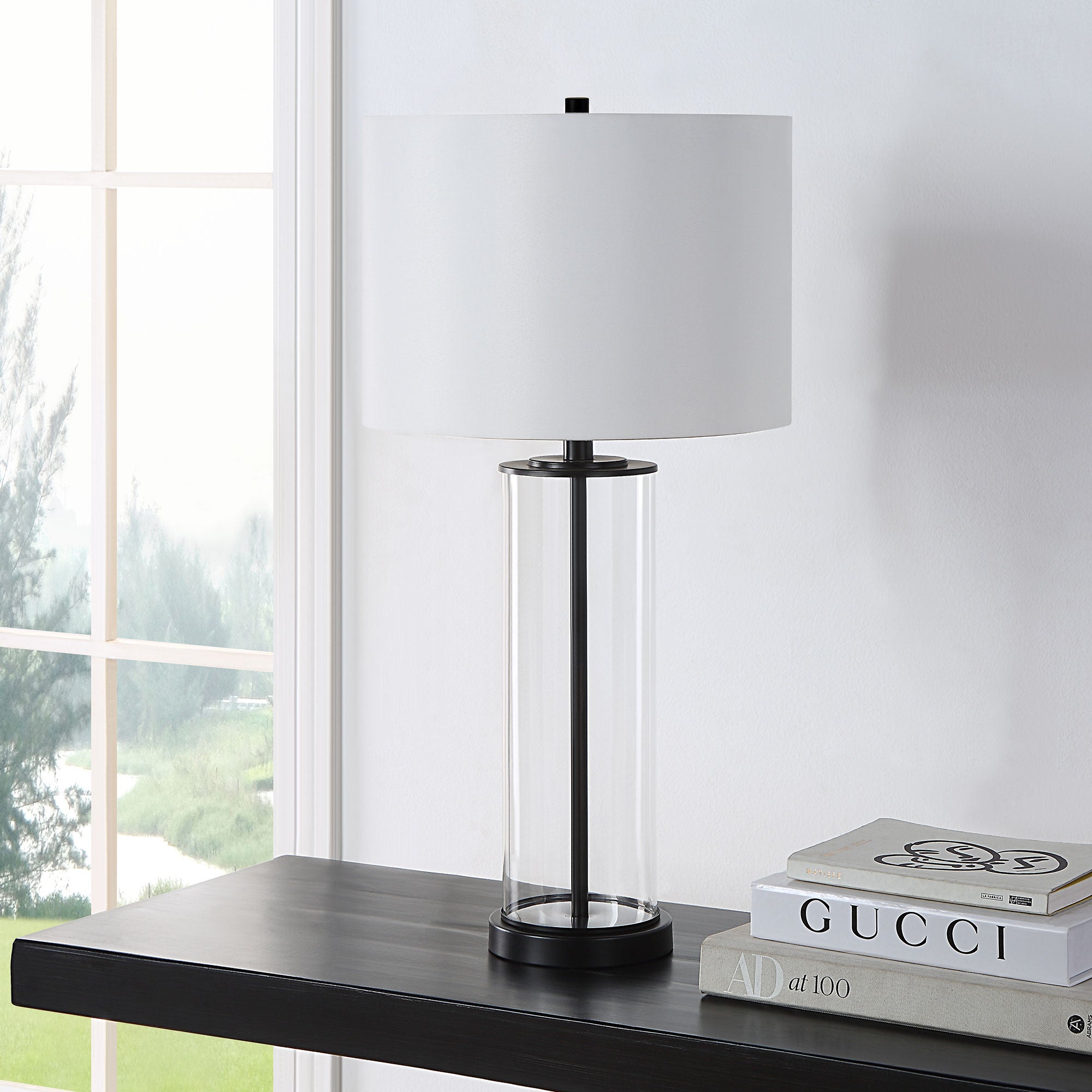 Desdemona Table Lamp placed on a shelf, featuring a clear glass base and white shade, near a window.