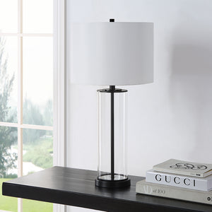 Desdemona Table Lamp placed on a shelf, featuring a clear glass base and white shade, near a window.
