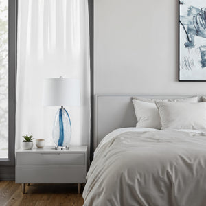 Solis Table Lamp on a nightstand in a modern bedroom, featuring a blue glass base and white shade.