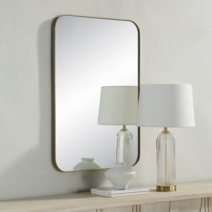 Vesey Wall Mirror with a rounded gold frame reflecting a lamp and vase on a wooden surface.