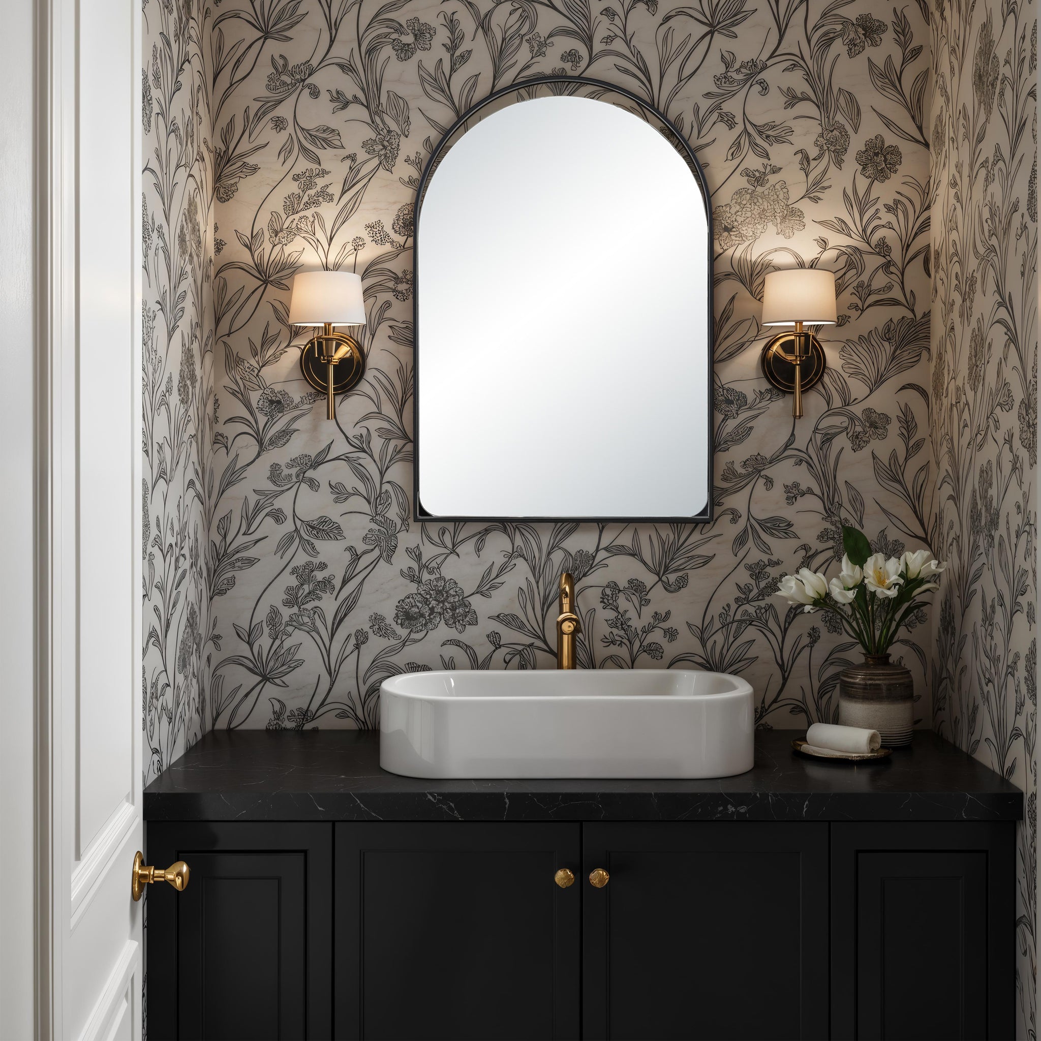 Maniola mirror with an arched design, mounted above a black countertop with a white basin, floral wallpaper, and gold accents.