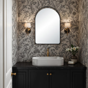 Maniola mirror with an arched design, mounted above a black countertop with a white basin, floral wallpaper, and gold accents.
